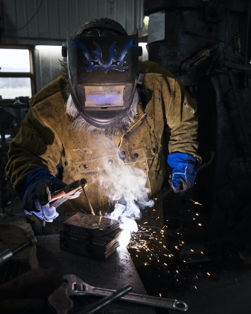 A welder wearing a protective mask and brown coat uses a torch, producing bright sparks and smoke in a dim workshop. The scene conveys focus and craftsmanship.
