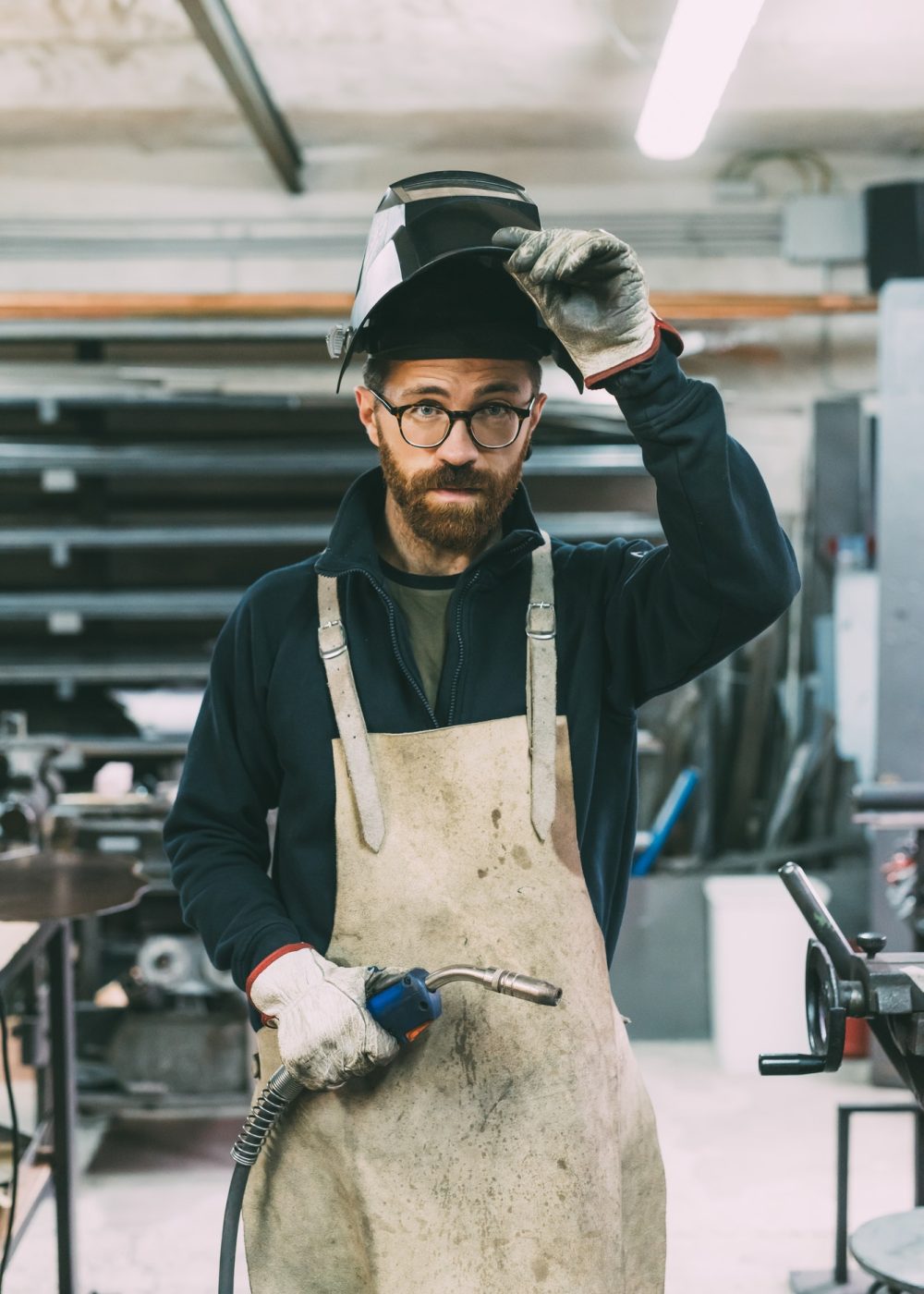 A craftsman in a workshop lifts his welding mask, revealing glasses and a thoughtful expression. He wears a dusty apron and holds welding tools. Shelving and equipment are in the background.