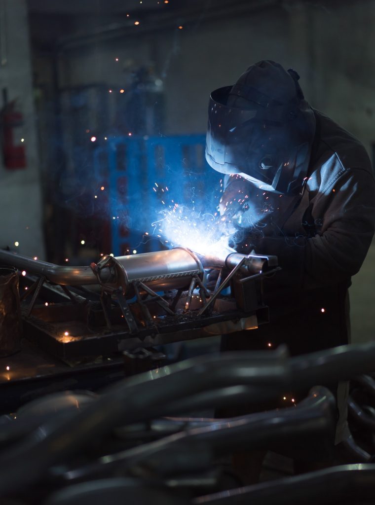 A welder in a dark workshop is engaged in welding metal pipes, emitting bright blue sparks. The scene conveys focus and industrial craftsmanship.