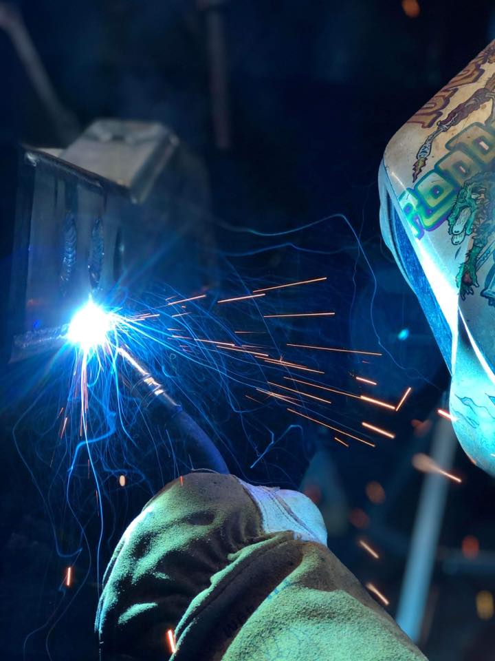 A welder operates a torch, emitting bright blue light and sparks, with a gloved hand. The scene conveys intense focus and skilled craftsmanship.