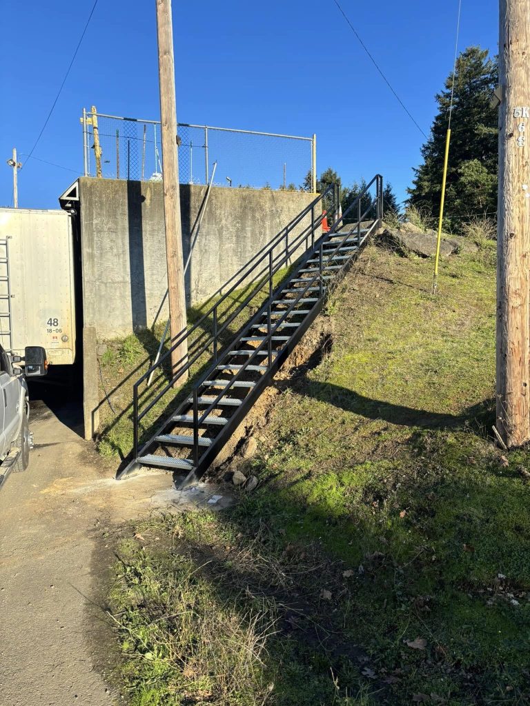 A steep metal staircase ascends a grassy hill alongside a concrete structure and wooden poles under a clear blue sky, creating an industrial yet serene vibe.
