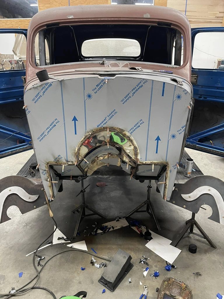 A partially restored vintage car body on a stand in a workshop. The metalwork shows welding and new panels. Tools and scattered materials lie on the floor.